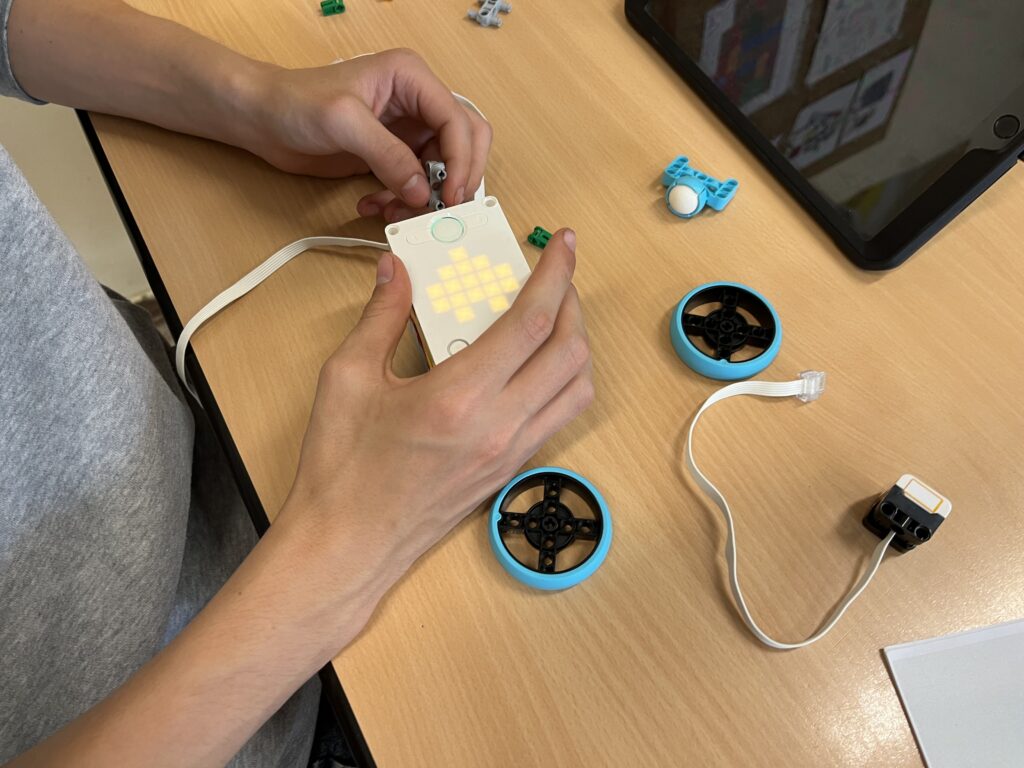 Hände montieren ein Bauteil an einem Lernroboter mit leuchtender LED-Matrix; daneben liegen Räder, Sensor und Tablet.